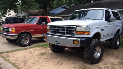 1994 Ford Bronco - Two Ford Broncos, a white lifted model and a red one, are parked side-by-side in a residential area.