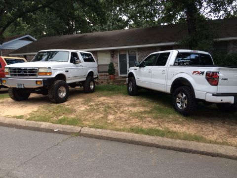 1994 Ford Bronco - Two white trucks, a lifted Ford Bronco and a Ford F-150, are parked in front of a suburban house.