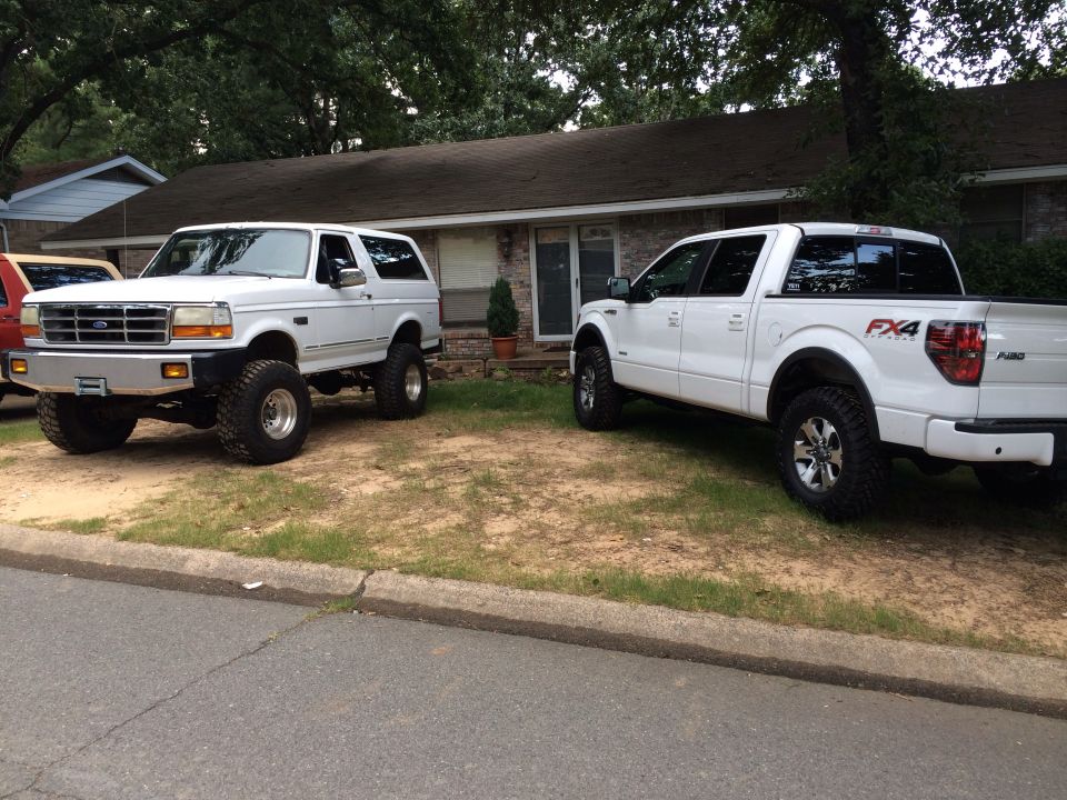 White Ford Bronco and a white Ford F-150 parked in a driveway. Bronco has aftermarket bumper and lift kit