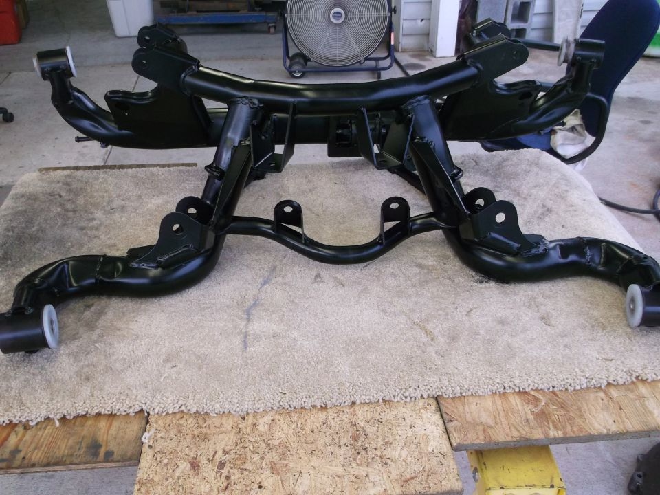 Black metal suspension subframe with white bushings, resting on a rug and wood planks
