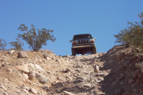 1992 Ford Bronco - Dark Ford Bronco cresting a rocky desert hillclimb, shot from below against a clear blue sky so the front grille, chrome bumper and exposed…