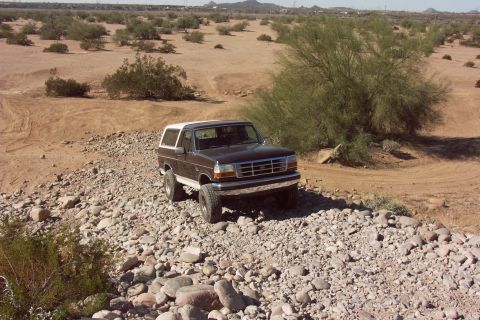 1992 Ford Bronco - Elevated three-quarter front view of a dark brown lifted Ford Bronco with white hardtop picking its way across a rocky dry wash strewn with…