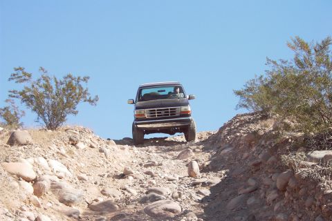 1992 Ford Bronco - Dark lifted Ford Bronco with a driver at the wheel poised at the top of a steep, rock-strewn desert descent, seen head-on from below with t…
