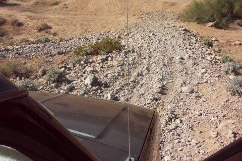 1992 Ford Bronco - Driver's-eye view over the dark hood and fender of a truck with a whip antenna, looking down a rocky wash strewn with river cobble in the d…