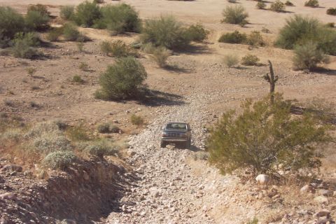 1992 Ford Bronco - Distant head-on view of a dark 1990s Ford Bronco picking its way up a rocky, cobble-filled desert wash. Sun-bleached sand, creosote brush a…