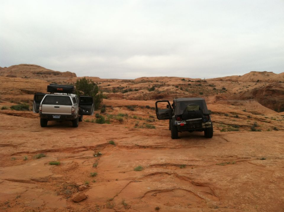 Two off-road vehicles, a silver pickup truck with a cap and a dark Jeep Wrangler, are parked on a desert trail with doors open