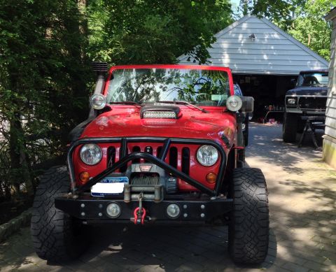 1979 Ford Bronco - Modified red Jeep Wrangler in front of a garage, with another vehicle visible behind it.