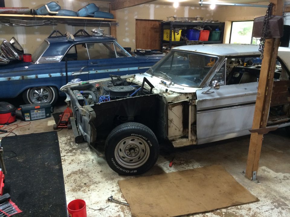Two old cars, one white and partially disassembled, the other blue, sit in a garage with parts on shelves above