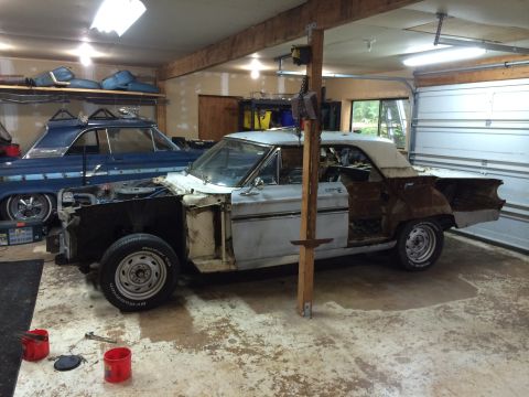 1964 Ford Fairlane - Two classic cars are in a garage, one undergoing restoration with its front end removed.