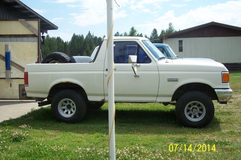 1990 Ford Bronco - A white 1990 Ford Bronco is parked on a grassy lawn next to a building, with another vehicle visible behind it.