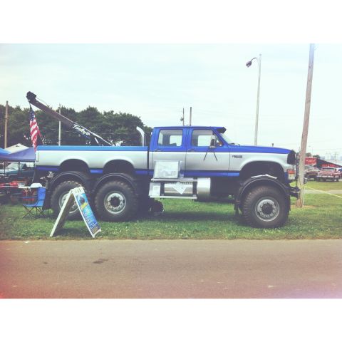 1979 Ford Bronco - A custom six-wheeled truck with a tow boom is parked on a grassy area next to a road.