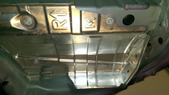 Underside of a vehicle showing metal shielding with 'R1' and 'M' stamped on it. There's a pipe and a black rubber plug visible