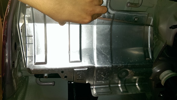 Close-up of a metal panel on a vehicle's undercarriage, possibly near the hitch area. A hand is visible reaching towards it