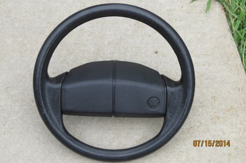 1985 Volkswagen Jetta - A black Volkswagen steering wheel is shown lying on a concrete surface with grass in the background.