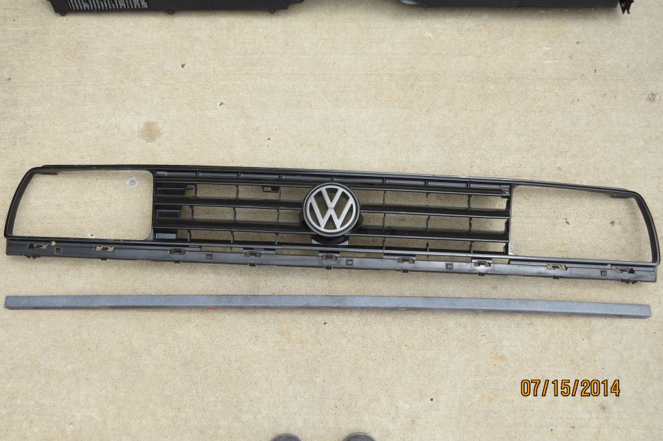 Black Volkswagen grille with silver emblem, horizontal bars, and rectangular headlight openings. A gray trim piece lies below