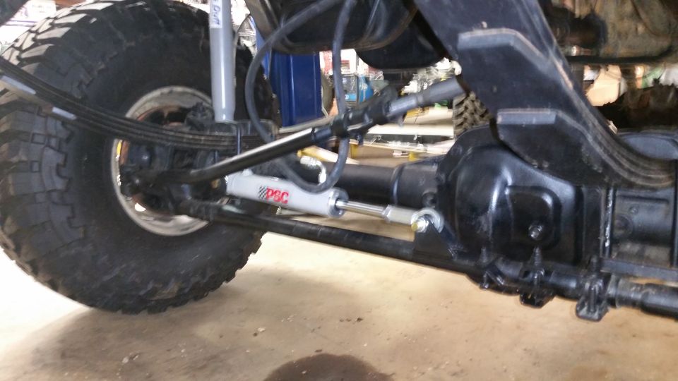 Close-up of a vehicle's rear axle, leaf springs, shock absorber, and steering stabilizer with 'PSC' logo