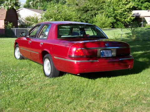 1999 Mercury Grand Marquis - A red 1999 Mercury Grand Marquis is parked on a grassy lawn with houses and trees in the background.