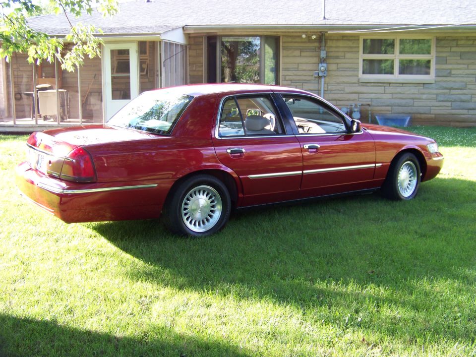 Red sedan parked on grass in front of a house. The car appears to be a Mercury Grand Marquis, but the model year is likely earlier than 1999