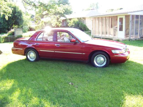 1999 Mercury Grand Marquis - A red sedan sits on a grassy lawn in front of a house, viewed from the side.