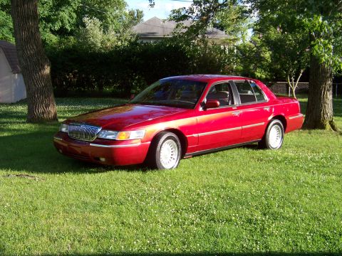1999 Mercury Grand Marquis - A red sedan is parked on a grassy lawn with trees and a house in the background.
