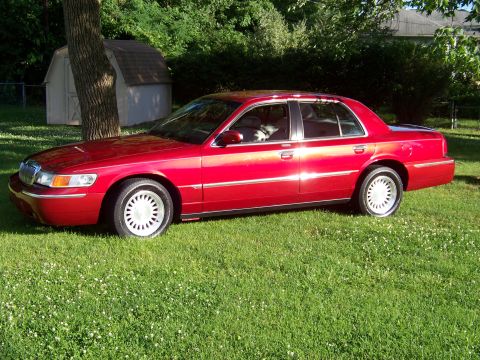 1999 Mercury Grand Marquis - A red 1999 Mercury Grand Marquis sedan is parked on a grassy lawn with a tree and shed in the background.