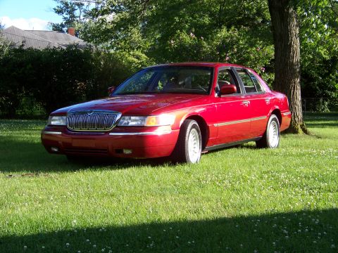 1999 Mercury Grand Marquis - A red 1999 Mercury Grand Marquis is parked on a grassy lawn next to a tree.