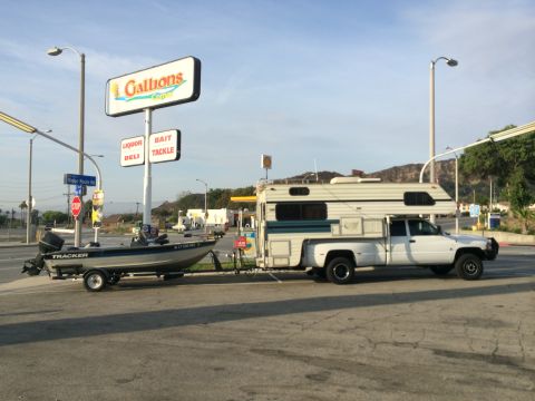 1997 Dodge 3500 - A white Dodge pickup truck tows a boat trailer while carrying a camper shell, parked near a gas station.