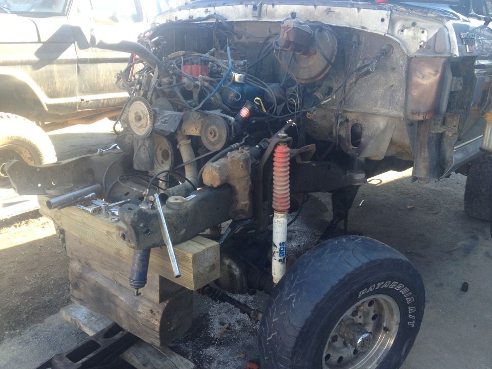 1979 Ford Bronco chassis disassembled, engine exposed, supported by wooden blocks. Another vehicle is partially visible to the left