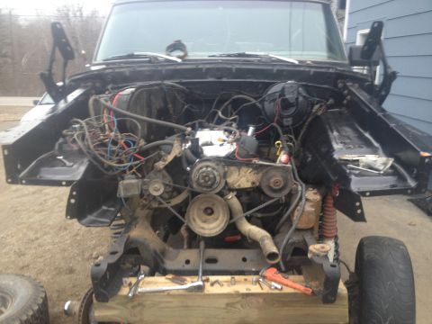 Vehicle engine bay with hood open, showing engine and wiring. Some tools are on a wooden support