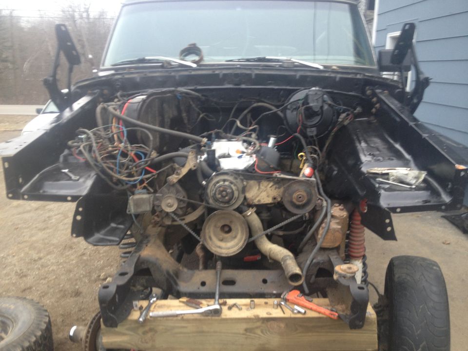 Vehicle engine bay with hood open, showing engine and wiring. Some tools are on a wooden support