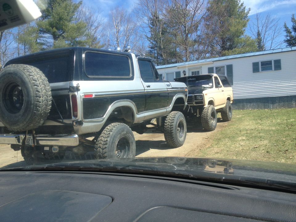 Two lifted Ford Broncos parked in front of a mobile home. The rear Bronco is black and silver with a spare tire on the back. The front Bronco is tan