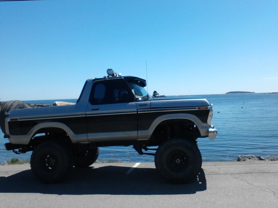 Lifted two-tone Ford Bronco parked by the water. Has a spare tire on the back and lights on the roof
