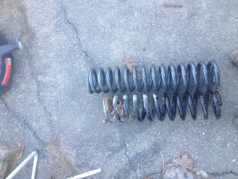 Two coil springs, one new and one rusted, lie on a concrete surface next to a black oil container