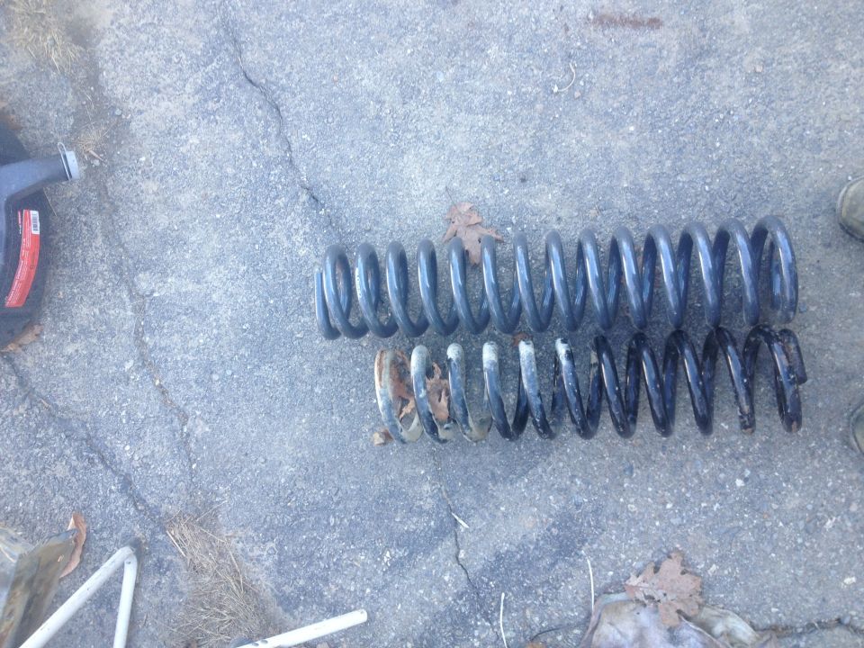 Two coil springs, one new and one rusted, lie on a concrete surface next to a black oil container