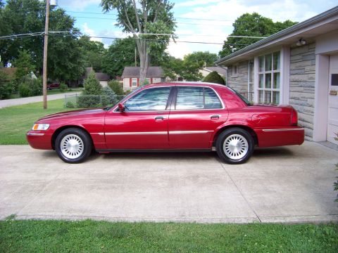 1999 Mercury Grand Marquis - A red 1999 Mercury Grand Marquis is parked on a concrete driveway next to a brick house.