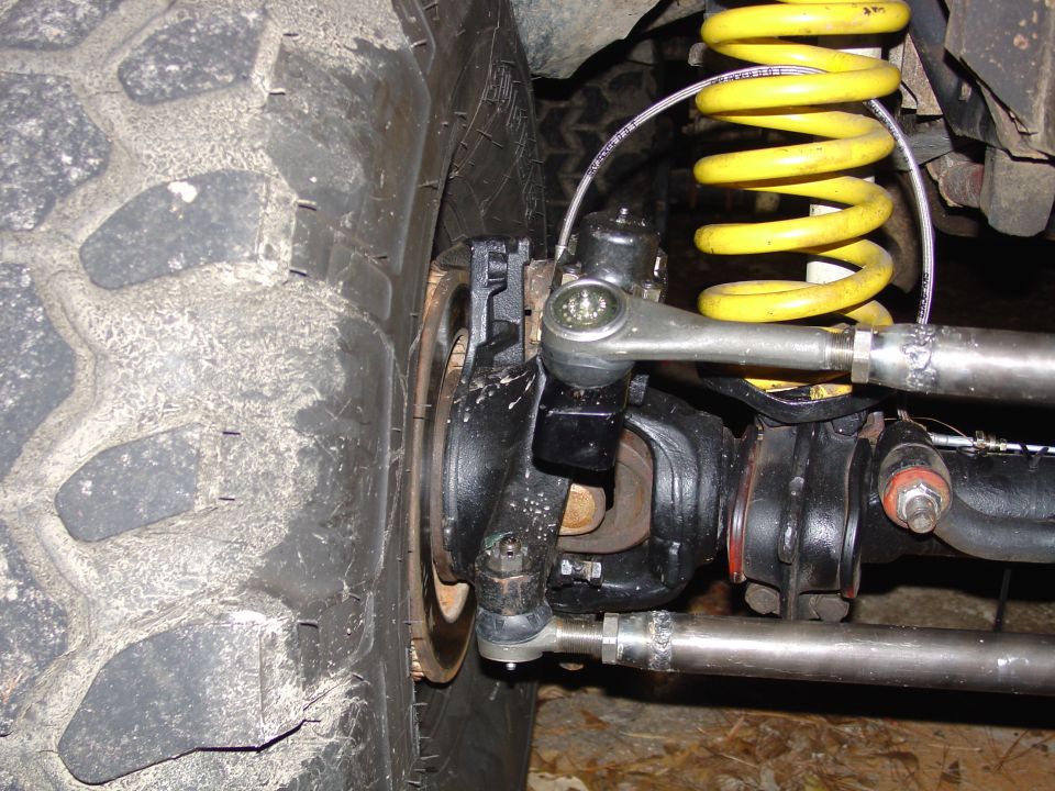 Close-up of a vehicle's front wheel, steering linkage, and yellow coil spring