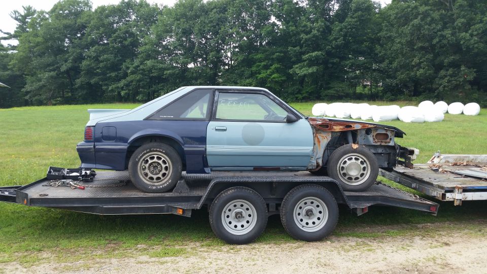 Two-tone blue Ford Mustang coupe on a trailer, missing front end and showing rust