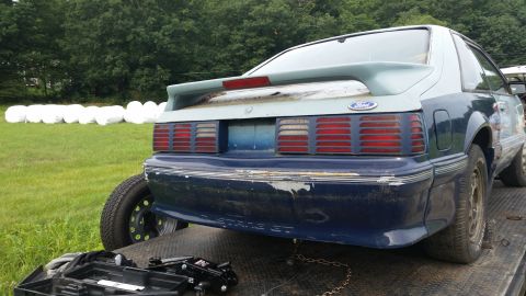 1991 Ford Crown Victoria LTD - The rear of a blue 1991 Ford Mustang GT is visible, loaded onto a tow truck with a field of wrapped bales in the background.
