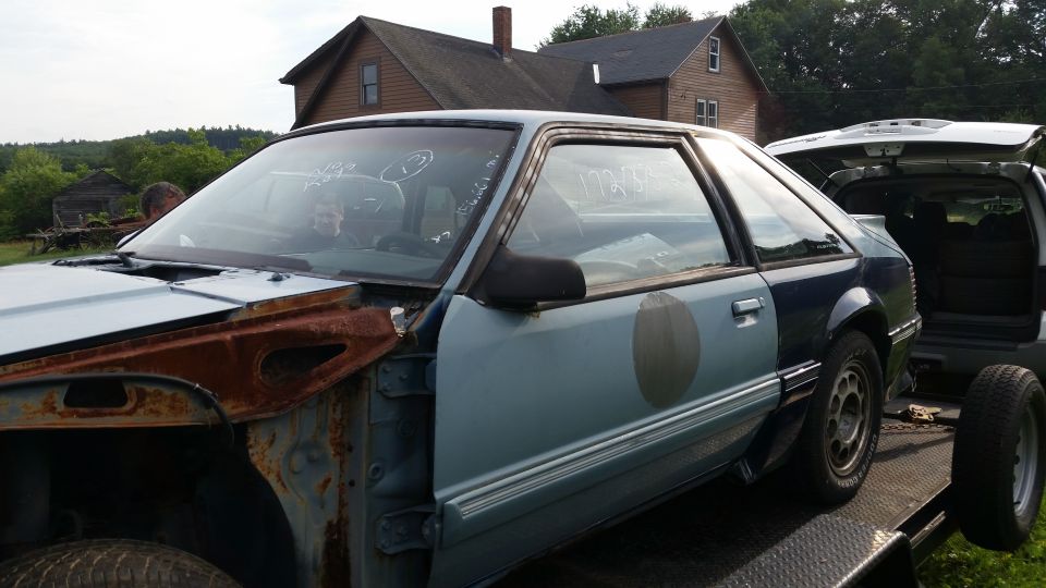 Light blue Ford Mustang coupe on a trailer, front fender removed, rust visible
