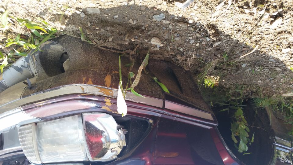 front corner of a dark red car, partially covered by dirt and vegetation. Headlight and trim are visible