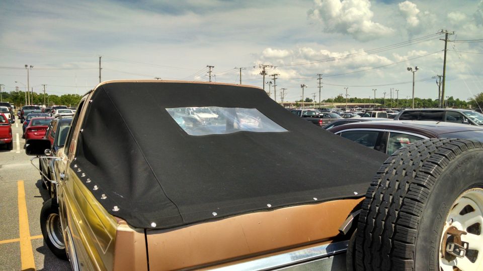 Rear view of a tan Ford Bronco with a black soft top and a spare tire mounted on the back. Other cars are in the parking lot