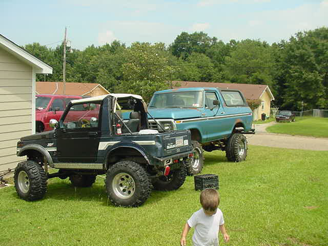 Blue 1978 Ford Bronco with lifted suspension and large tires parked on grass next to a black off-road vehicle
