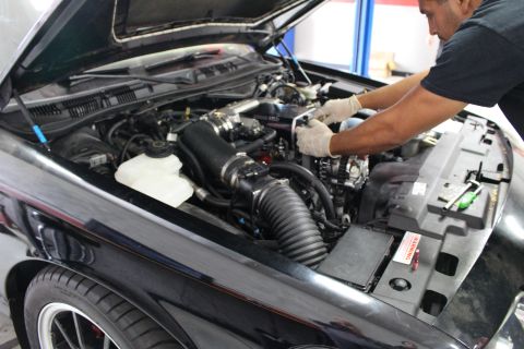 2003 Ford Crown Victoria - gloved mechanic works in the engine bay of a black car fitted with black corrugated intake tubing and a supercharger.