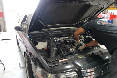 2003 Ford Crown Victoria - black Ford sedan with its hood raised in a shop, a mechanic reaching into the engine bay over the front fender.