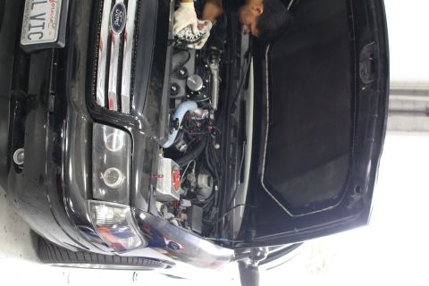 2003 Ford Crown Victoria - mechanic works on the supercharged engine of a black Ford sedan showing the Ford grille badge and a custom plate.