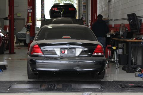 2003 Ford Crown Victoria - Rear view of a black Ford sedan strapped on a dynamometer inside a busy tuning shop with lifts.