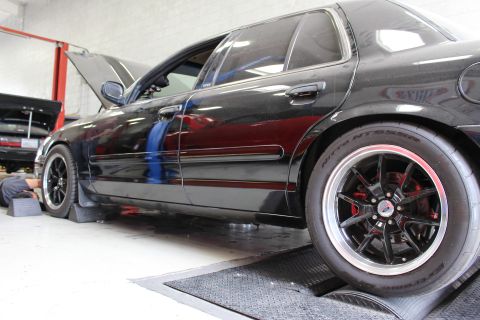 2003 Ford Crown Victoria - Side profile of a black Ford sedan with black multi-spoke wheels and Nitto tires parked on a shop dyno.