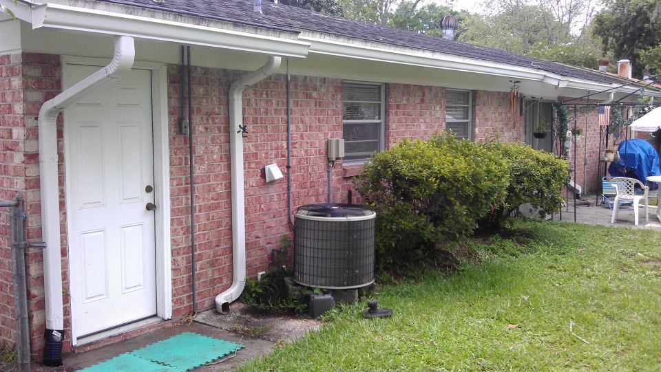 side of a brick house with a white door, downspouts, and an outdoor AC unit. Some bushes and grass are visible