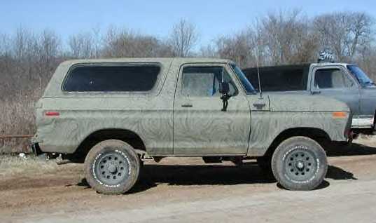 Side view of a 1978 Ford Bronco in camouflage paint, parked next to another vehicle