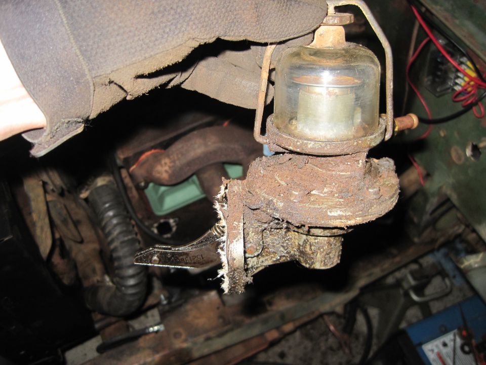 Close-up of a rusty fuel pump with glass sediment bowl, held by a gloved hand, with engine exhaust manifold visible